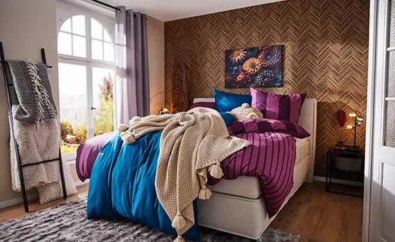 Gemütliches Schlafzimmer mit großem Bett, gestreiften Kissen, Decken und Teppich. Fenster mit Blick nach draußen, Leiter mit Decken.