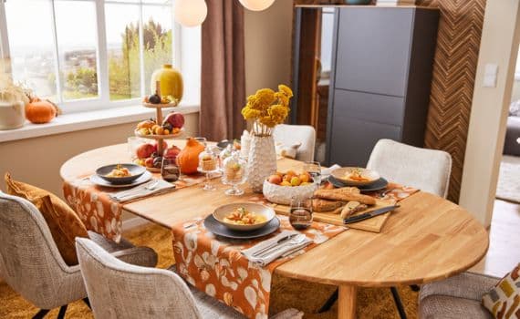 Herbstlich gedeckter Esstisch mit vier Gedecken, orangefarbenen Tischläufern, Kürbissen, Blumen und Obstschale in hellem Esszimmer.
