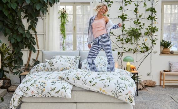 Frau in gestreifter Pyjamahose und rosa Top tanzt auf einem Bett mit floralem Bettbezug in hellem, begrüntem Raum.
