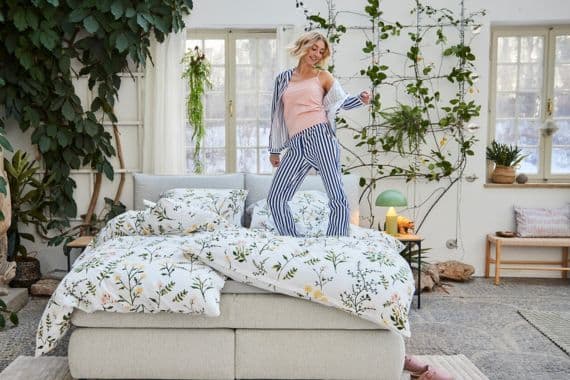 Frau in gestreifter Pyjamahose und rosa Top tanzt auf einem Bett mit floralem Bettbezug in hellem, begrüntem Raum.