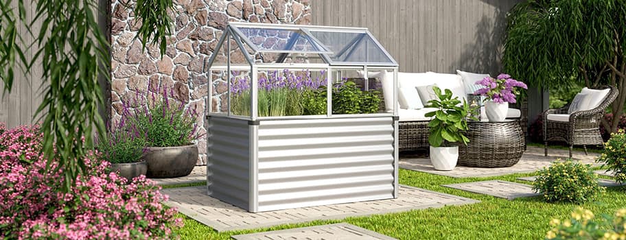 Kleines Gewächshaus im Garten mit blühenden Pflanzen, daneben Gartenmöbel vor Steinmauer.