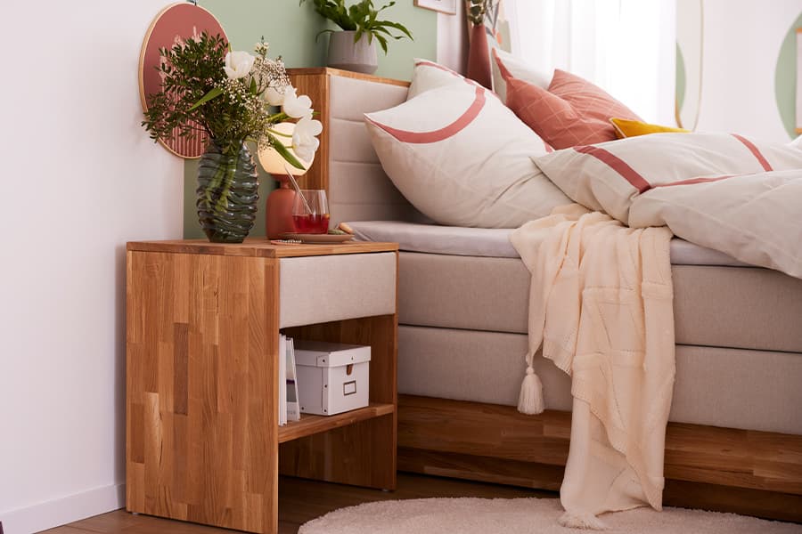 Nachttisch aus Holz neben einem beige gepolsterten Bett mit weißer Bettwäsche und dekorativen Kissen. Auf dem Nachttisch stehen eine Vase mit Blumen und eine Lampe. Eine beige Decke hängt über dem Bett.