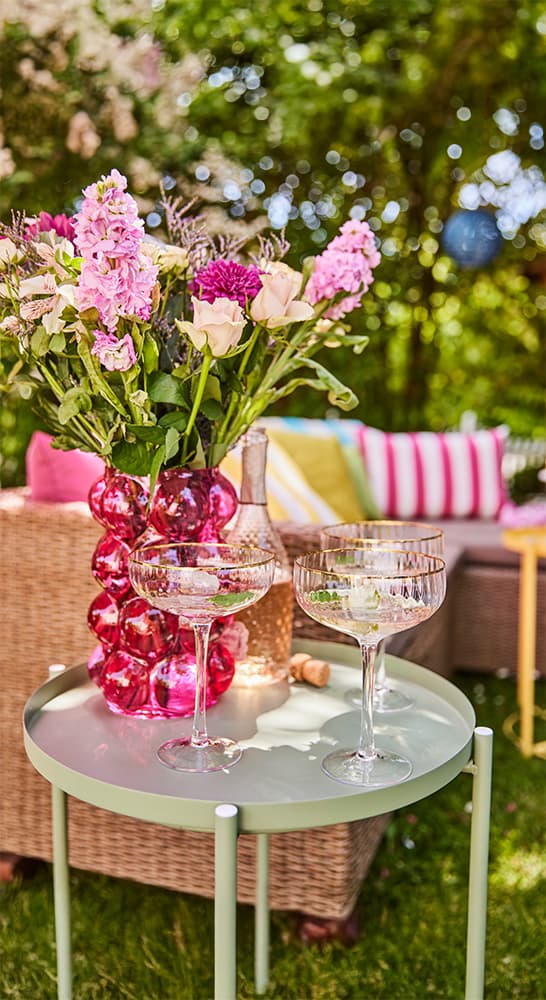 Vase mit bunten Blumen auf grünem Tisch, zwei Gläser und Flasche daneben, im Hintergrund Gartenmöbel mit Kissen