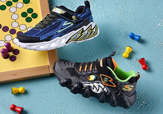 Zwei Paar Kinderturnschuhe – eines in Blau mit Blitzmuster, das andere in Schwarz mit Monsteraugen – neben Brettspiel und Spielfiguren