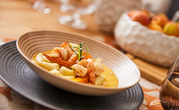 Teller mit Garnelen und Pasta in gelber Soße, dekoriert mit Rosmarin, im Hintergrund unscharfe Schale mit Obst
