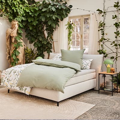 Gemütliches Schlafzimmer mit grün-weißer Bettwäsche, Pflanzen und Statue neben Fenster.
