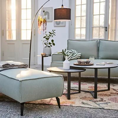 Wohnzimmer mit Sofa und Hocker, runden Couchtischen, Stehlampe, Pflanzen und großen Fenstern im Hintergrund