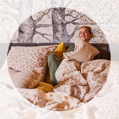 Frau sitzt lachend im Bett, umgeben von gemusterter Bettwäsche und Kissen vor einer Wand mit Baum-Motiv