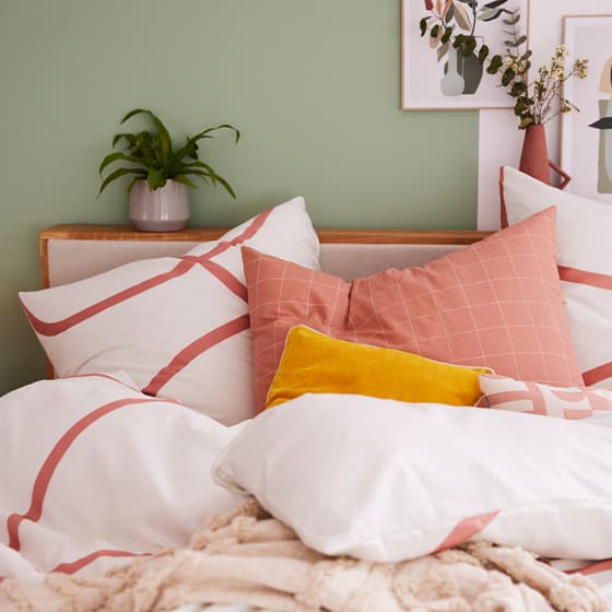 Bett mit weißer Bettwäsche, roten Kreismustern und farbigen Kissen vor einer grünen Wand mit Pflanzen und Bildern.