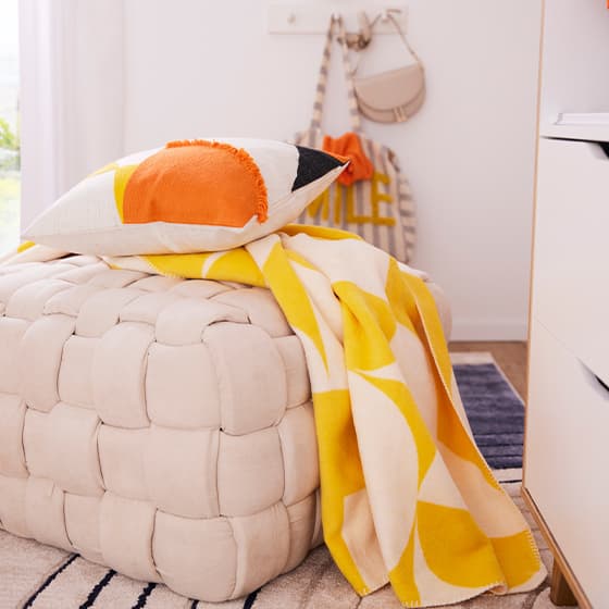 Beiger gepolsterter Sitzhocker mit einem dekorativen Kissen und einer gelb-weißen Decke, im Hintergrund eine Tasche und ein Hut an der Wand.