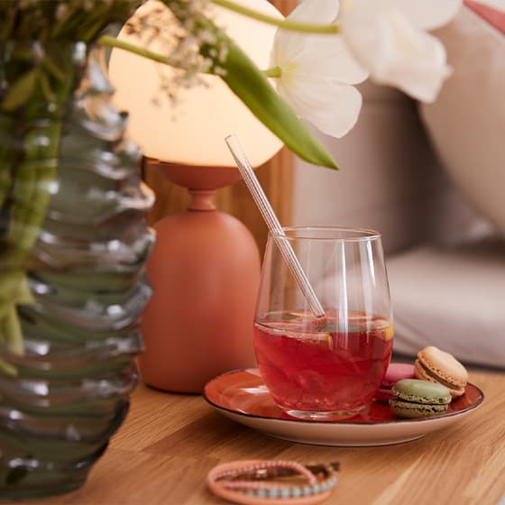 Ein Glas mit rotem Getränk und Strohhalm auf einem Teller, begleitet von Macarons. Im Hintergrund eine Tischlampe und eine Vase mit Blumen.