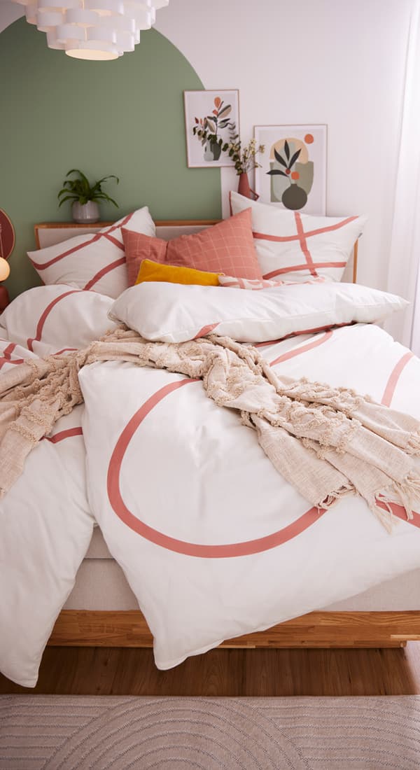 Bett mit weißer Bettwäsche und roten Kreismustern, dekoriert mit verschiedenen Kissen in Rot, Gelb und Weiß. Eine beigefarbene Decke liegt über dem Bett. Im Hintergrund eine grün gestrichene Wand mit gerahmten Bildern und Pflanzen.