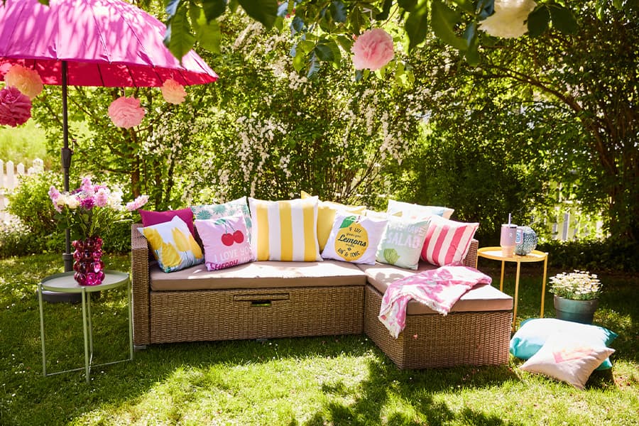 Sommerlicher Garten mit gemütlichem Sofa, bunten Kissen, pinkem Sonnenschirm, Blumenstrauß und Beistelltischen auf grünem Rasen