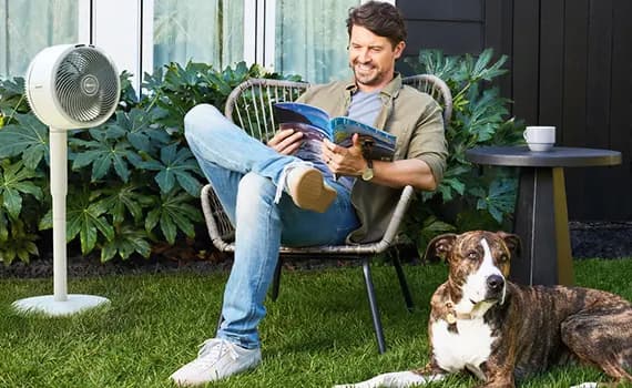 Mann liest Buch auf Gartenstuhl neben Ventilator, Hund liegt im Gras, Tisch mit Tasse im Hintergrund