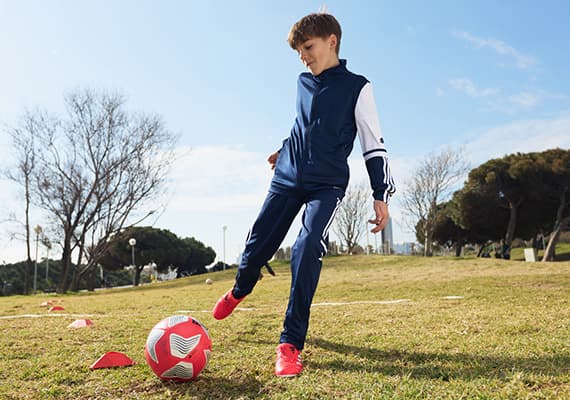 Junge in blau-weißer Sportkleidung kickt roten Fußball auf grüner Wiese mit Bäumen im Hintergrund