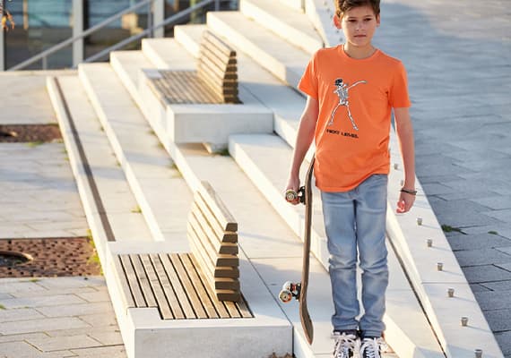 Junge in orangefarbenem T-Shirt mit Skateboard-Motiv hält Skateboard, steht auf Treppenstufen neben Holzbank