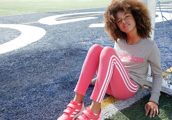 Mädchen mit rosa Adidas-Leggings und grauem Adidas-Pullover sitzt auf Sportplatz, Netz im Hintergrund