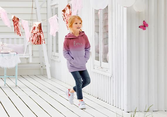 Kind mit rosa-lila Pullover mit "love" Schriftzug, Jeans und weißen Sneakers auf Holzterrasse vor weißem Haus.