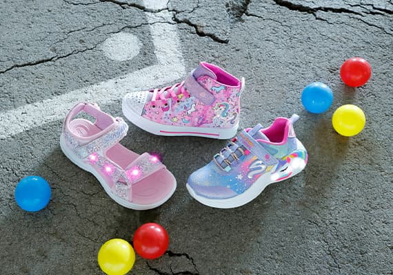 Drei Paar Kinder-Schuhe: pinke Sandalen, hohe pinke Sneaker mit Einhörnern, lilafarbene Sneaker mit bunten Mustern. Bunte Bälle drumherum.