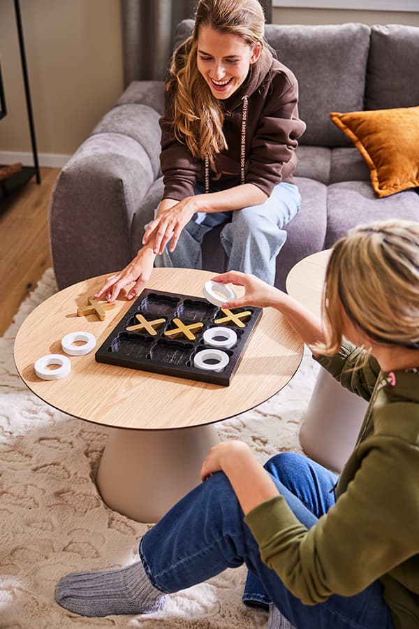 Zwei Personen spielen Tic-Tac-Toe auf einem runden Tisch im Wohnzimmer, eine sitzt auf dem Sofa, die andere auf dem Teppich.