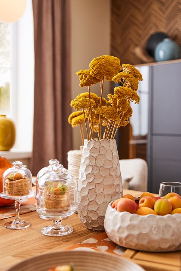 Esstisch mit Trockenblumen in einer Vase, Aprikosen in einer Schale und Kuchenstücken unter Glasglocken in einem gemütlichen Raum
