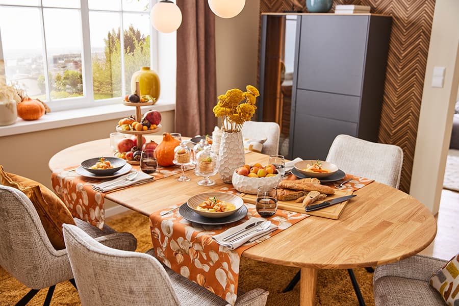 Festlich gedeckter Esstisch mit Herbstdekoration, darunter Kürbisse und Blumen. Teller mit Speisen und Brotkorb auf dem Tisch.