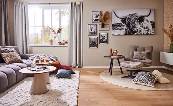 Gemütliches Wohnzimmer mit grauem Sofa, rundem Tisch, Dekokissen und Sesseln. Wandbilder und ein großes Fenster im Hintergrund.