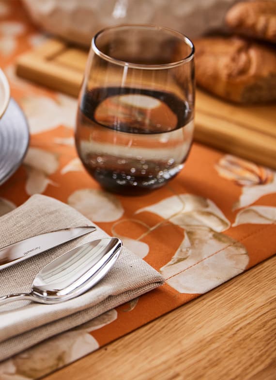 Glas Wasser auf orangefarbenem Blumen-Tischläufer neben Besteck und Stoffserviette, dahinter Brot auf Holzbrett