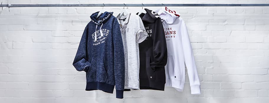 Vier Kapuzenpullover und ein T-Shirt hängen nebeneinander auf einer Stange vor weißer Ziegelwand
