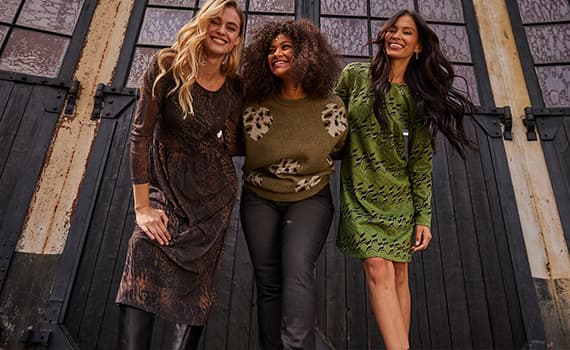 Drei Frauen in herbstlicher Kleidung, zwei in Kleidern und eine in Pullover und Hose, stehen vor einer Holztür.