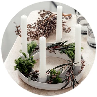 Weißer Adventskranz mit vier weißen Kerzen, dekoriert mit grünen Zweigen und braunen Beeren, steht auf einem Tisch.