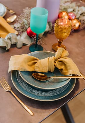 Festlich gedeckter Tisch mit blauem Teller, darauf gelbe Serviette in Schleifenform, goldene Gabel und bunte Gläser.