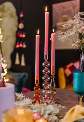 Drei brennende Kerzen in unterschiedlichen Kerzenständern auf einem Tisch, umgeben von Dekorationen und Blumen.