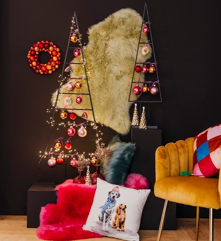 Weihnachtsdeko mit zwei Metallbäumen, roten Kugeln, Lichterketten, Fell, Kissen mit Hundemotiv und gelbem Sessel vor dunkler Wand