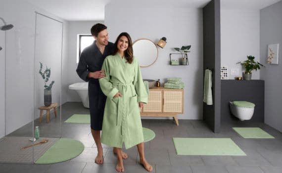 Paar in Bademänteln, Mann in dunkelblau, Frau in hellgrün, steht in modernem Badezimmer mit grünen Teppichen und Pflanzen