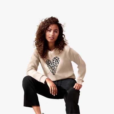 Frau mit lockigem Haar trägt cremefarbenen Pullover mit Herzmotiv aus Leopardenmuster und schwarze Hose