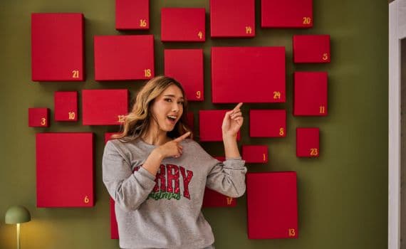 Eine Frau steht vor einer olivgrünen Wand, an der ein selbstgemachter Adventskalender hängt, der aus roten Schachteln besteht, die mit goldenen Zahlen von 1 bis 24 beschriftet sind.