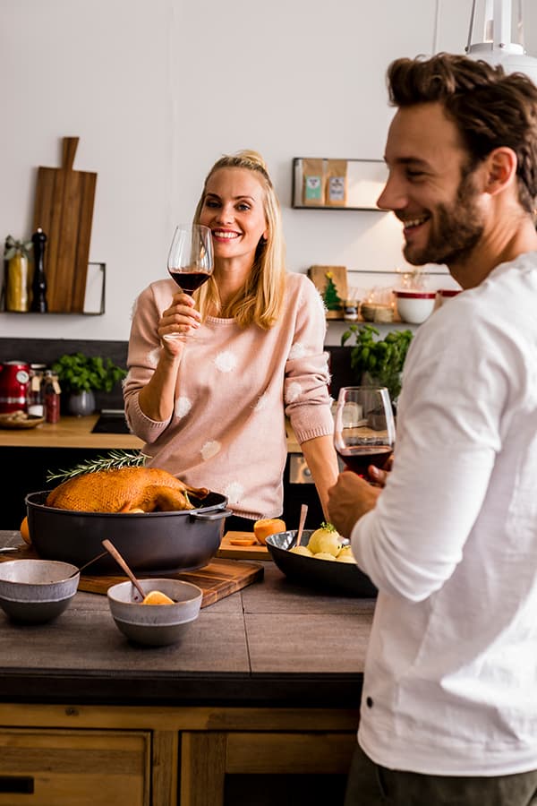 Zwei Personen genießen Wein in einer Küche, ein Braten und Schalen mit Gemüse stehen auf dem Tisch.