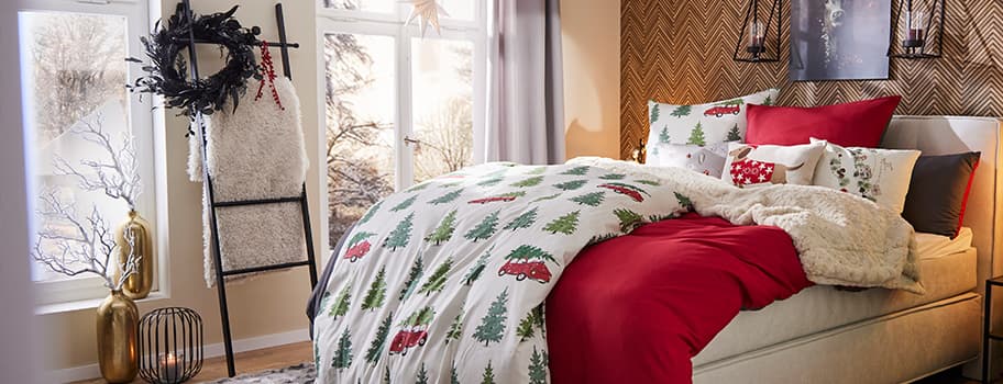 Weihnachtlich dekoriertes Schlafzimmer mit Bettwäsche mit Tannenbäumen und roten Autos, roter Bettdecke und Kissen mit Sternen.