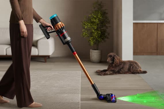 Person saugt Boden mit einem kabellosen Staubsauger, Hund liegt auf Holzfußboden neben einem grünen Laserlicht