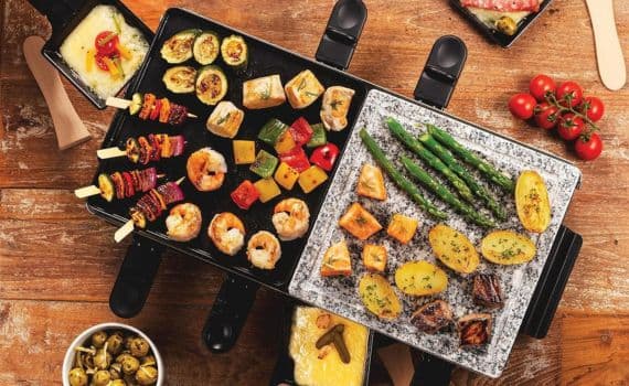 Verschiedene gegrillte Speisen auf Raclette-Grill, darunter Gemüse, Kartoffeln, Fisch und Garnelen.