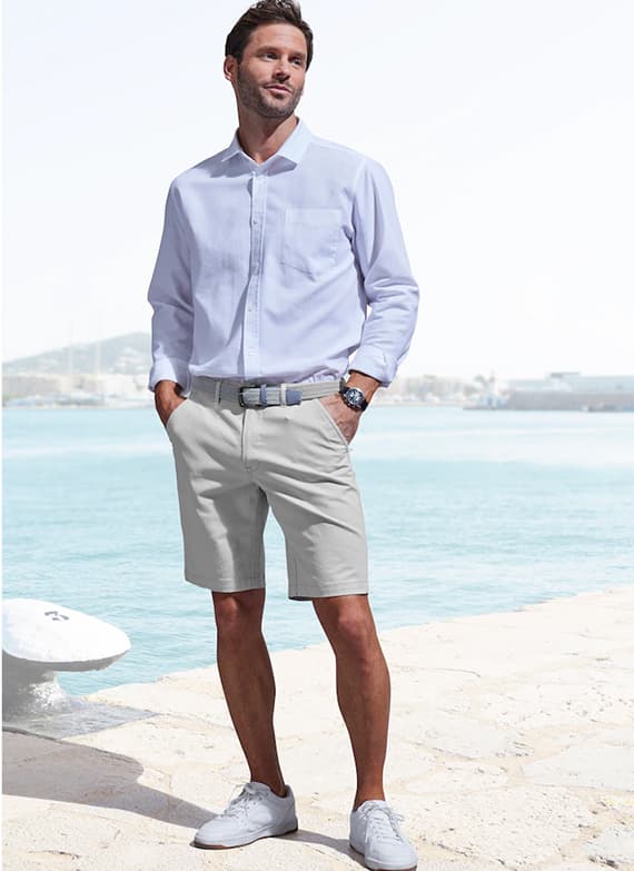 Mann in hellen Shorts und weißem Hemd steht mit Händen in den Taschen am Hafen vor dem Meer
