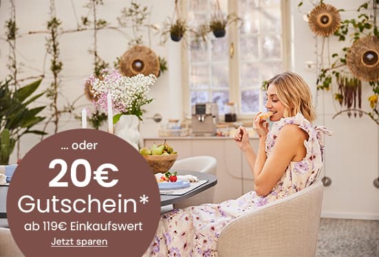 20€ Gutschein ab 119€ Einkaufswert – Frau im geblümten Kleid sitzt am Esstisch und isst Obst in hellem Raum