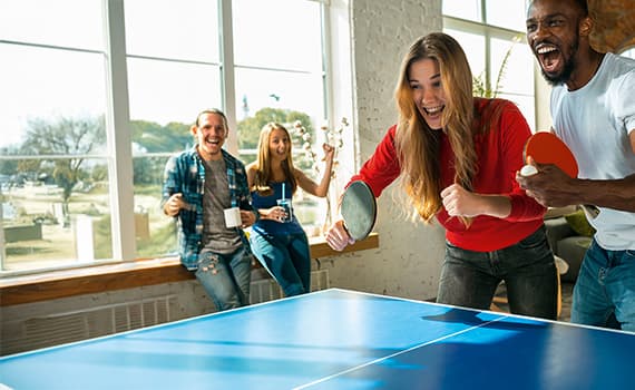 Frau in rotem Shirt spielt Tischtennis mit Mann, zwei Personen sitzen im Hintergrund mit Getränken