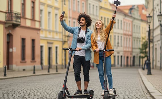 Zwei Personen auf E-Scootern machen Selfies in einer malerischen Altstadt mit bunten Gebäuden im Hintergrund
