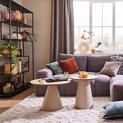 Gemütliches Wohnzimmer mit grauem Sofa, bunten Kissen und zwei runden Beistelltischen. Regal mit Pflanzen und Deko rechts im Hintergrund.