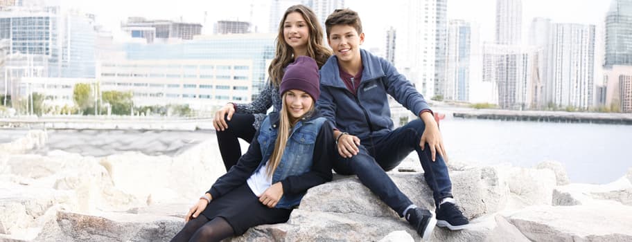 Drei Kinder sitzen auf Felsen am Wasser, im Hintergrund moderne Gebäude und ein Hafen.