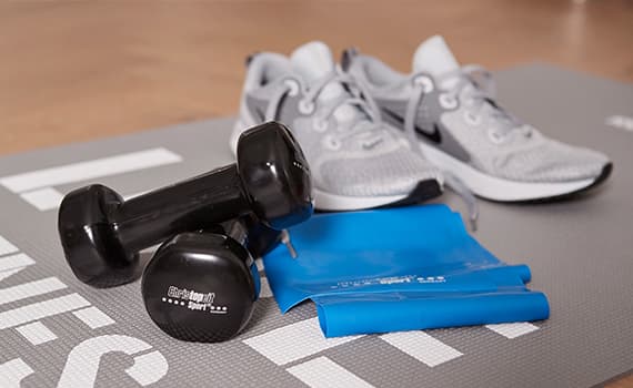 Schwarze Hanteln, blaues Widerstandsband und graue Sportschuhe auf einer Fitnessmatte mit weißen Schriftzügen