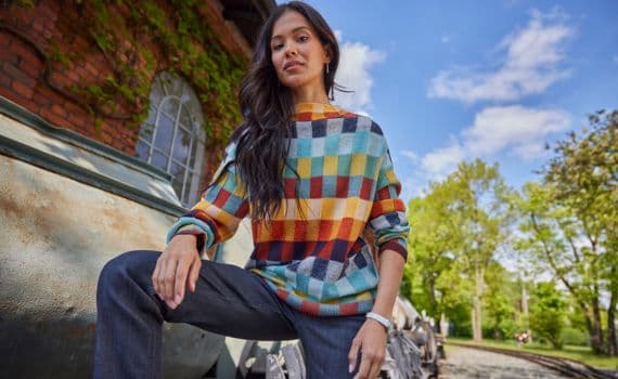 Frau in farbigem Karopullover und dunkler Hose vor einem Gebäude im Freien.