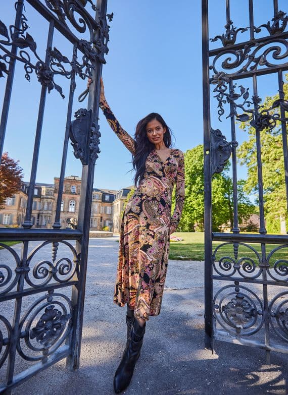 Frau in gemustertem Kleid und schwarzen Stiefeln steht in einem offenen Tor, im Hintergrund ein historisches Gebäude und Bäume.